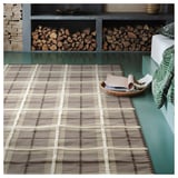 A brown and grey wool rug with a classic tartan check pattern, displayed in a room with a green bench and stacked firewood.