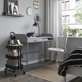 Small black mobile storage trolley beside modern gray desk in office. Holds headphones, books, and box. Fits under desk for extra space.