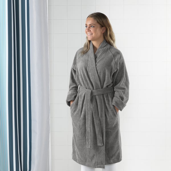 Woman wearing gray ROCKÅN bathrobe with adjustable belt, side pockets, and cuffed sleeves, standing against tiled wall.