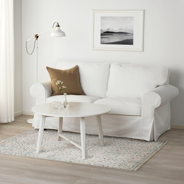 Living room with white floral rug, sofa, coffee table, vase, beach art, lamp.
