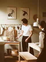 Two people cook in a modern kitchen. A man organizes dishes by a trolley, a woman arranges plates at a table.