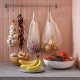 Two bamboo bowls hold fruits and veggies on a countertop. Above, net bags hang with produce against peach-tiled walls.