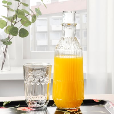 Clear glass carafe with ornate design, containing orange liquid, sitting on a tray, accompanied by a matching glass.
