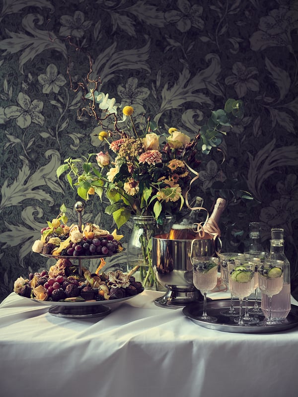 Elegant table setup with SÄLLSKAPLIG glassware, fresh flowers, and fruits on a white tablecloth, set against dark floral wallpaper.
