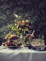 Elegant table setup with SÄLLSKAPLIG glassware, fresh flowers, and fruits on a white tablecloth, set against dark floral wallpaper.