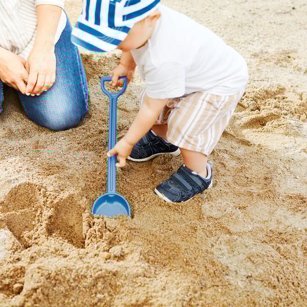 A child plays with a blue sand shovel on a sandy surface, likely at a beach, with an adult nearby.