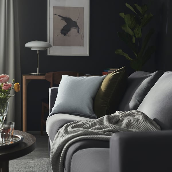Cozy living room with gray couch, colorful pillows, dark walls, plant, art, lamp, side table with flowers and drink.