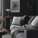 Cozy living room with gray couch, colorful pillows, dark walls, plant, art, lamp, side table with flowers and drink.