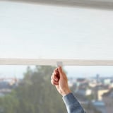 A hand adjusts a light filtering roller blind, suitable for tall windows, enhancing privacy while allowing soft light.