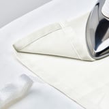 A silver iron rests on off-white SANELA curtains laid out flat, ready for pressing.