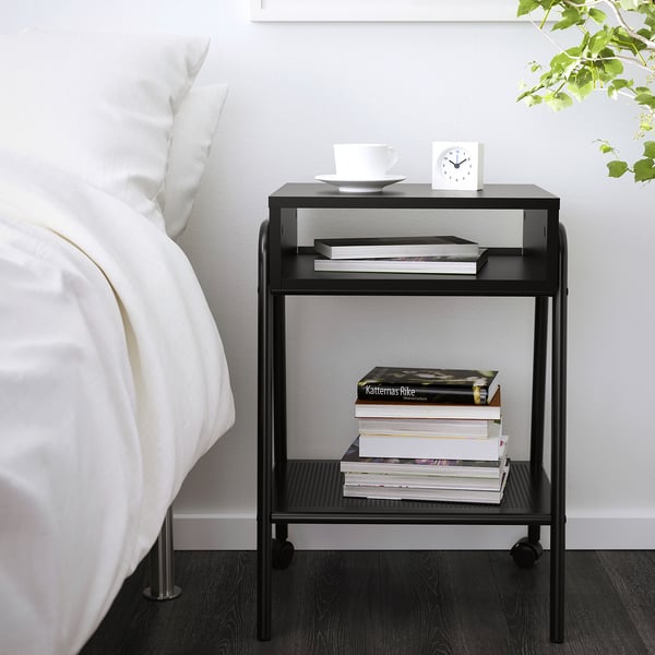 Small black metal nightstand beside a bed with books, white bowl, and alarm clock.