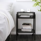 Small black metal nightstand beside a bed with books, white bowl, and alarm clock.