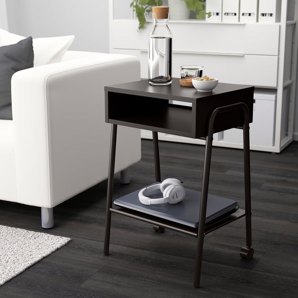Modern black SETSKOG bedside table with wheels, perfect for a compact space next to a white sofa, holding a laptop and headphones.