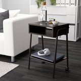Modern black SETSKOG bedside table with wheels, perfect for a compact space next to a white sofa, holding a laptop and headphones.