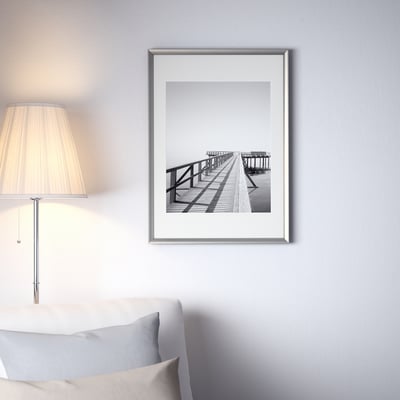 Black and white a wooden dock on a wall. Large silver frame above a bed with a lamp and a pillow.