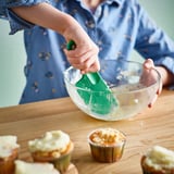 Person mixes batter with green spatula in glass bowl on wooden table. Frosted muffins nearby.