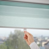 Hand adjusts cordless roller blind, ensuring safe, soft closure, filtering daylight.