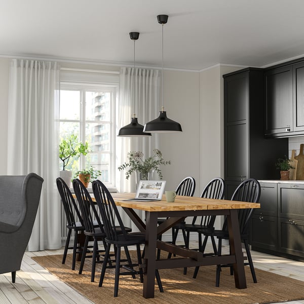 SKOGSTA 6-seat dining set with acacia table & spindle-back chairs, near window with lamps & plants.