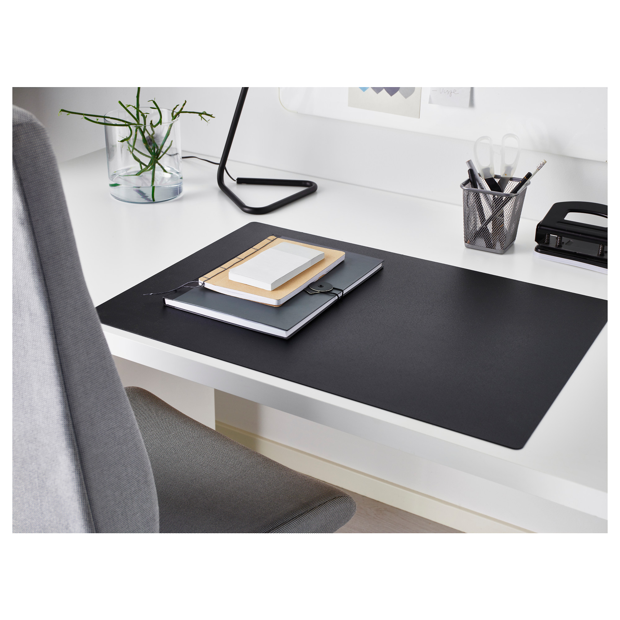 Modern workspace with black SKRUTT desk pad, notebooks, pen holder, and small plant. Pad covers desk, has smooth surface.