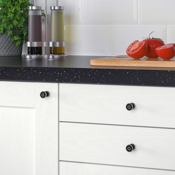 Two tomatoes on a cutting board, flanked by bottles with dispensers, set on a black speckled countertop against a white tiled backsplash.