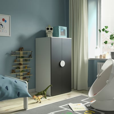 Childs room with gray wardrobe, blue car pillow bed, toy shelf, green curtains, white rocker, dinosaur toy.