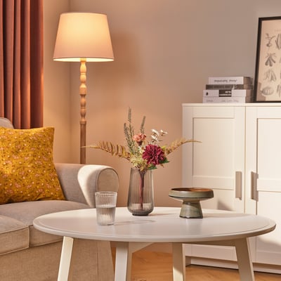 A living rooms coffee table displays a glass vase with artificial flowers and a glass tumbler beside a lamp.