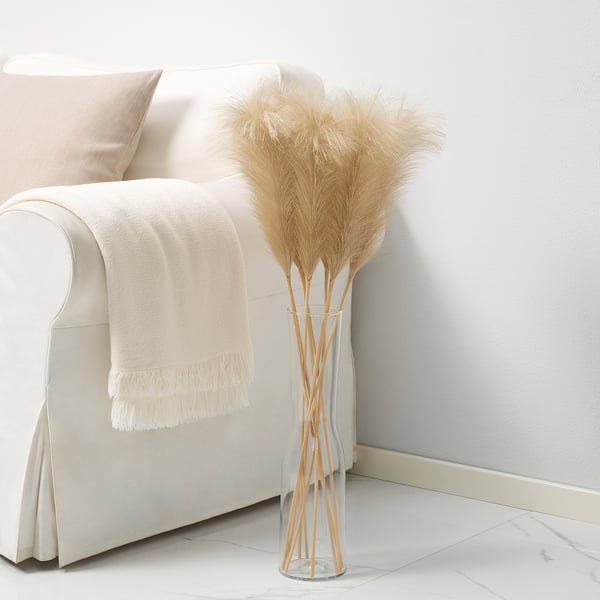 SMYCKA pampas grass in clear vase. Tall, beige plumes bendable at stems. White sofa with cream blanket and pillow. Modern, minimalist design.