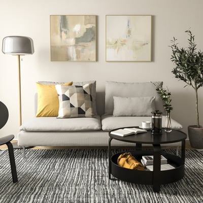 Modern living room with SÖDERHAMN sofa, white with patterned pillows, round black coffee table, floor lamp, and abstract art on walls.