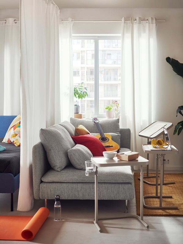 Bright living space: gray SÖDERHAMN sofa, red pillows, guitar, window with white curtains, plants, and small tables.