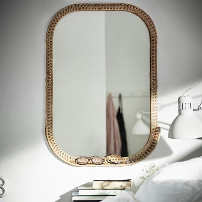 Large, woven natural rattan mirror on wall, reflecting a white lamp, clothes, and partially showing a bed, adding cozy, warm feel.