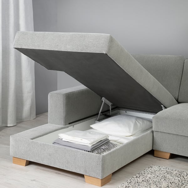 Modern sofa with storage ottoman open, showing blankets inside. Light grey fabric, wooden legs.