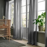 Modern room with STENFRÖ sheer gray curtains, large potted plant, wicker chair. Curtains soften light, puddle on floor, framed by tall windows.