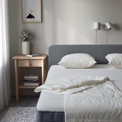 Minimalist bedroom with white bed, grey headboard, STJÄRNSTARR duvet, nightstand with flowers, books, lamp. Serene decor.