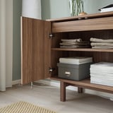 Wooden cabinet in walnut veneer with open door revealing neatly stacked towels, papers, and a storage box.