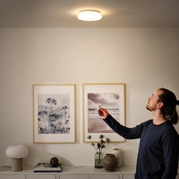 Man dims wall lamp with remote in modern living room featuring two framed pictures and decorative vases on a sideboard.