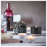 Cozy table setup with lit STÖRTSKÖN candles. Candles in black, patterned, and clear holders. Soft glow on a wooden table.