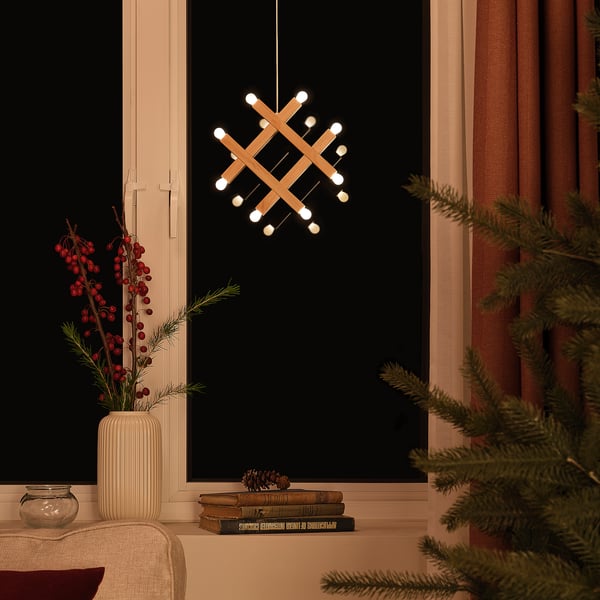 Modern hanging lamp with a pine wood design, illuminated with warm white lights, placed near a window, adorned with berry branches and pine cones.