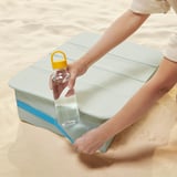 In a person is placing a water bottle on a blue and white beach tray table, demonstrating its functionality for outdoor use.