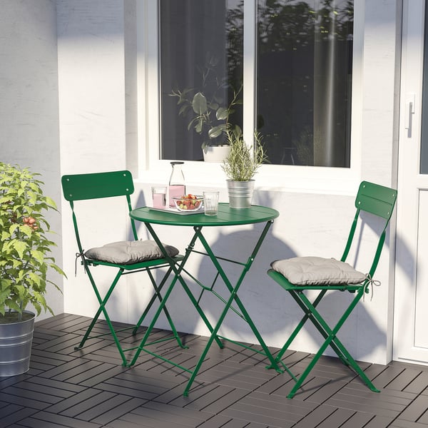 Small green metal bistro set with cushioned seats on balcony. Round table holds drinks & snacks. Suitable for compact spaces.