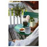 Small outdoor table in green, folded and ready for balcony. It has a round top and powder-coated steel frame, holding a drink, pastry, and flowers.