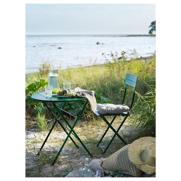 Green outdoor table and chair set by water, with drinks and hat.