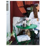 Woman enjoying drink at green SUNDSÖ outdoor table with white chair on balcony.