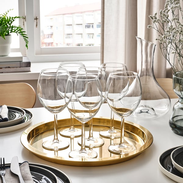 Six clear wine glasses on a golden tray alongside plates and cutlery, positioned near a window with white curtains.