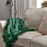 Green SVINROT throw blanket with white/grey patterns on beige sofa. 100% cotton, recycled, with lamp in background.