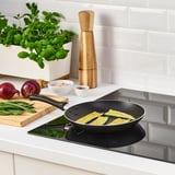 Black frying pan on induction hob with sliced zucchini cooking, surrounded by fresh vegetables and herbs on wooden cutting board.