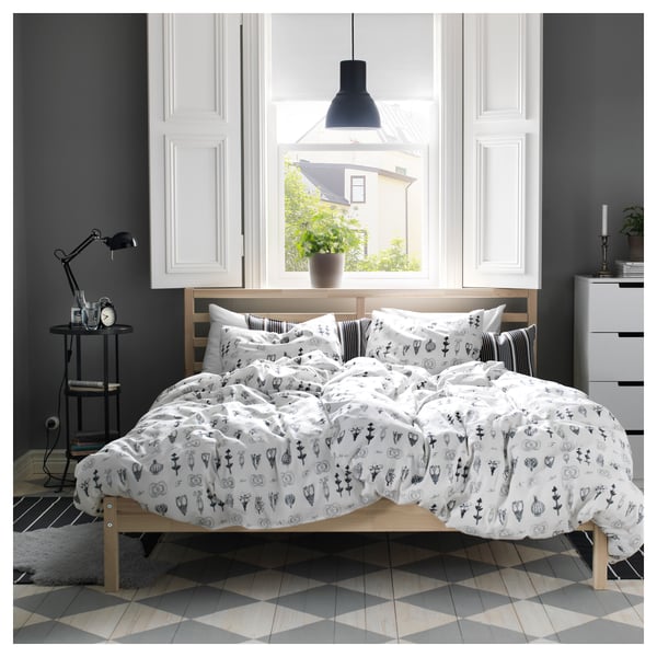 Modern wooden bed with white patterned bedding against grey wall. Natural wood frame with large black pendant lamp above, and grey tiled floor.