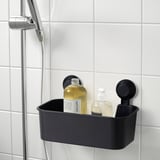 Small black TISKEN shower caddy with two compartments holds shampoo and soap next to chrome shower head on tiled wall.