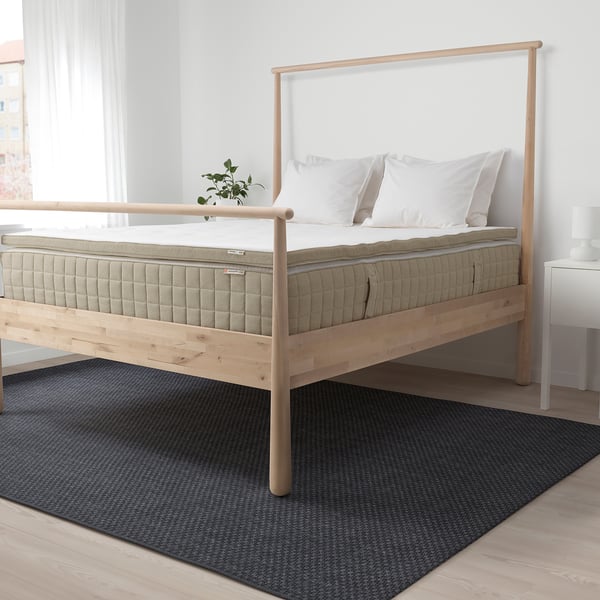 Modern wooden bed with large, natural-colored TISTEDAL latex mattress, two white pillows, small plant. Dark blue rug.