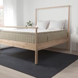Modern wooden bed with large, natural-colored TISTEDAL latex mattress, two white pillows, small plant. Dark blue rug.