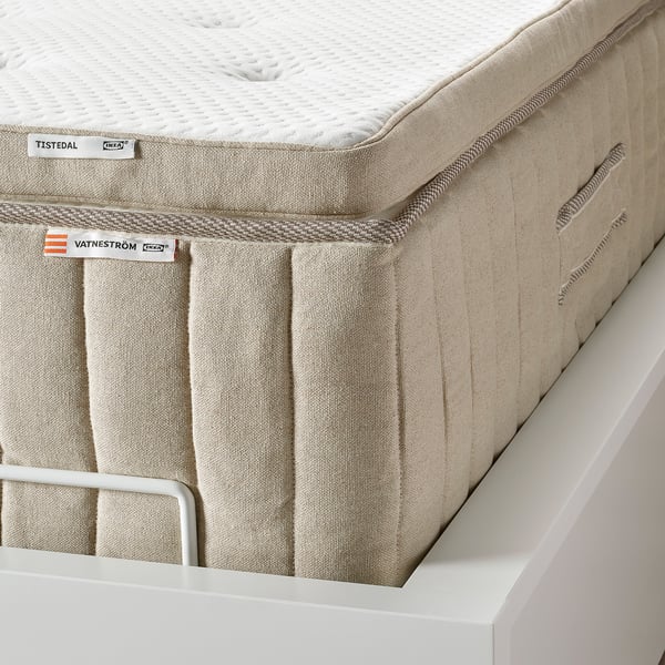 Image shows a corner of an IKEA TISTEDAL mattress pad on a bed frame, highlighting its breathable natural latex and soft, stretch fabric top layer.